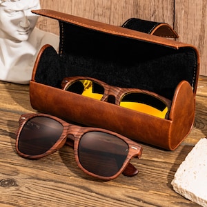 Puede incluir: Una funda de cuero marr&oacute;n con interior negro contiene dos pares de gafas de sol. Un par es marr&oacute;n con un patr&oacute;n de veta de madera y el otro par es amarillo con un marco dorado.