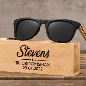 May include: Black sunglasses with wooden arms and a wooden box. The box is engraved with the text "Stevens JR. GROOMSMAN 29.04.2022".