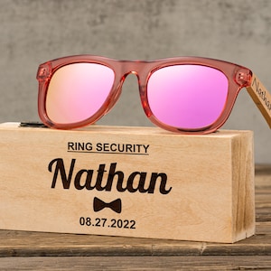 May include: A wooden box with a personalised inscription for "Nathan" and the date "08.27.2022". The box contains a pair of pink sunglasses with mirrored lenses. The sunglasses have a wooden frame.