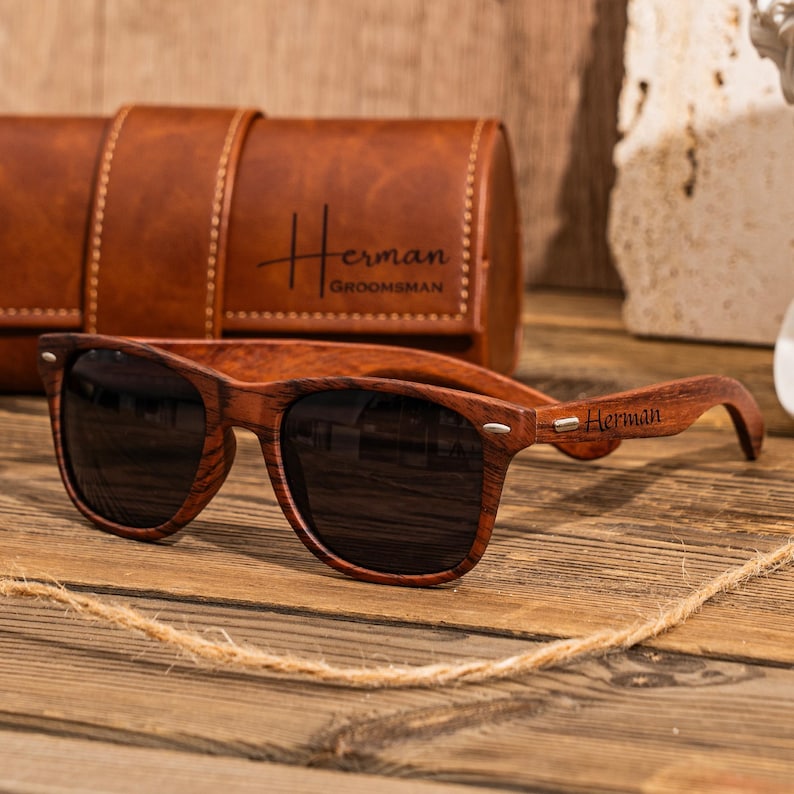 Puede incluir: Gafas de sol de madera con lentes oscuros y una funda de cuero marr&oacute;n. Las gafas tienen el nombre "Herman" grabado en el lateral. La funda tiene el nombre "Herman" y la palabra "Groomsman" grabada.