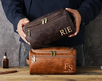 May include: Two rectangular leather toiletry bags with zippered closures and metal hardware. The top bag is dark brown with the monogram R|E. The bottom bag is tan with the monogram FS and the name FABIAN SCOTT printed below the initials.