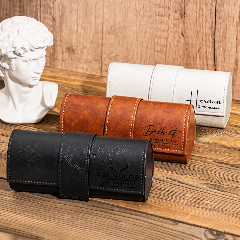 Puede incluir: Tres estuches para rollos de reloj personalizados en cuero negro, marr&oacute;n y blanco. Los estuches est&aacute;n enrollados y tienen una correa para mantenerlos cerrados. Los estuches est&aacute;n grabados con los nombres "Saunders", "Delbert" y "Herman" y el t&iacute;tulo "Groomsman".