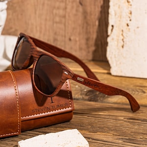 Puede incluir: Gafas de sol de madera roja con lentes marrones y una funda de cuero. Las gafas tienen el nombre "Steven" grabado en el lateral. La funda tiene el nombre "Steven Tumer" grabado.