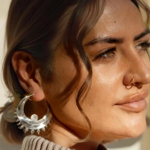 May include: Close-up of a person wearing large, silver hoop earrings. The earrings feature a crescent moon design with a central gem and dangling details. The person also has a gold nose ring.