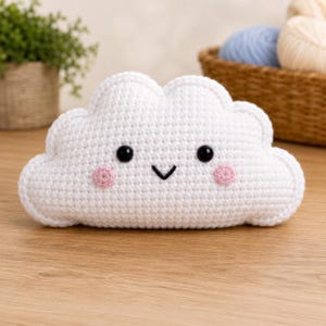 May include: A white cloud-shaped crochet plush toy featuring two black plastic safety eyes, a stitched black V-shaped mouth, and two small circular pink stitched cheeks. The texture is made of uniform white yarn stitches in a soft, rounded form.