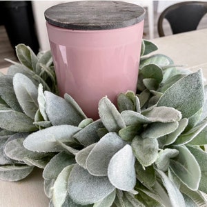 Pu&ograve; includere: Una candela rosa con un coperchio in legno grigio si trova in una ghirlanda di verde miller polveroso.