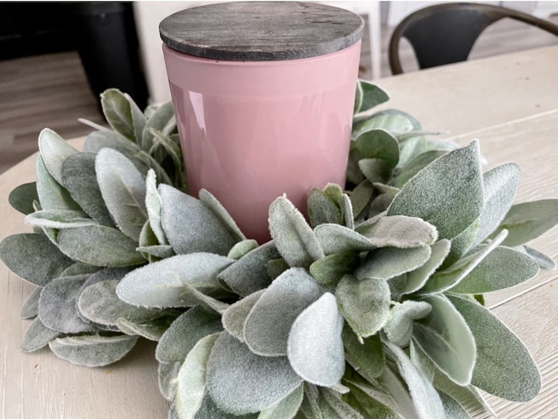 Pu&ograve; includere: Una candela rosa con un coperchio in legno grigio si trova in una ghirlanda di verde miller polveroso.