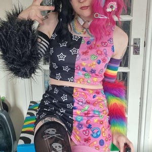 Könnte beinhalten: Ein zweiteiliges Outfit mit einem schwarz-weiß gemusterten Oberteil und Rock, kontrastiert durch einen rosa und bunten Druck. Das Ensemble umfasst Regenbogen-Armstulpen, Oberschenkel-Strümpfe und eine Perücke mit schwarzen und rosa Abschnitten.