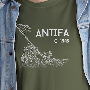 Puede incluir: Camiseta verde oliva con la palabra "ANTIFA" y "C. 1945" en texto blanco. Un gráfico blanco de soldados izando una bandera está debajo del texto. La camiseta se usa con una chaqueta vaquera azul.