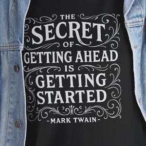Könnte beinhalten: Schwarzes T-Shirt mit dem Zitat "THE SECRET OF GETTING AHEAD IS GETTING STARTED" in weißer Schrift, zugeschrieben an Mark Twain. Das Shirt wird mit einer blauen Jeansjacke und Jeans getragen.