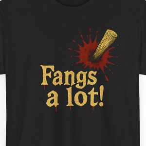 Könnte beinhalten: Schwarzes T-Shirt mit dem Schriftzug "Fangs a lot!" in Gold. Eine Grafik eines Holzpflocks, der einen roten Blutspritzer durchbohrt, befindet sich über dem Text. Das T-Shirt hat einen Rundhalsausschnitt.