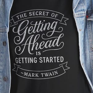 Könnte beinhalten: Schwarzes T-Shirt mit dem Zitat "THE SECRET OF Getting Ahead IS GETTING STARTED - MARK TWAIN" in weißer Schrift. Das Shirt wird mit einer blauen Jeansjacke und einer blauen Jeans getragen.