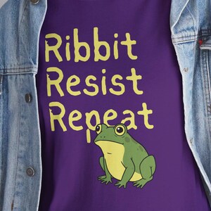 May include: Purple t-shirt with the words "Ribbit Resist Repeat" in yellow, and a cartoon frog illustration. The frog is green with a yellow belly. The shirt is worn with a denim jacket and blue jeans.