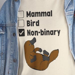 Könnte beinhalten: Cremefarbenes T-Shirt mit einer braunen Schnabeltier-Illustration und dem Text "Säugetier, Vogel, Non-binär". Das Schnabeltier liegt auf dem Rücken. Das Wort "Non-binär" ist angekreuzt. Die Person trägt eine Jeansjacke.