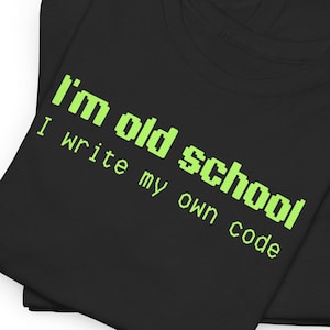 May include: Two black t-shirts are folded and stacked. The top shirt has the text "I'm old school I write my own code" in bright green, pixelated font. The shirts are made of a soft material.