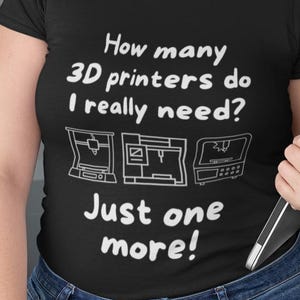 Op de afbeelding: Zwart t-shirt met de tekst "How many 3D printers do I really need? Just one more!" en drie printer illustraties. De persoon draagt een blauwe spijkerbroek en een bril.