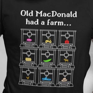 Op de afbeelding: Zwart T-shirt met de tekst "Old MacDonald had a farm..." boven een afbeelding van 3D-printers die elk een ander boerderijdier of object printen. Het shirt wordt gedragen door een persoon met een witte baard en tatoeages.