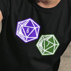 May include: Black t-shirt featuring two geometric dice designs. One die is purple with a white outline and the number 6, the other is green with a white outline and the number 7. The shirt is worn by a person wearing sunglasses and a gray cap.