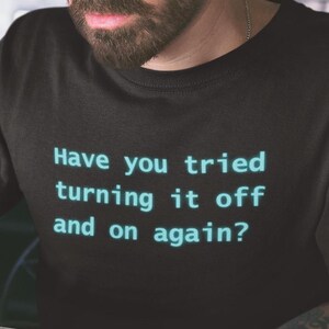 Może przedstawiać: Czarny t-shirt z napisem "Have you tried turning it off and on again?" w kolorze jasnoniebieskim. Osoba nosząca koszulkę używa laptopa. Na stole stoi szklanka z płynem.