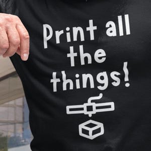 Op de afbeelding: Zwart T-shirt met de witte tekst "Print all the things!" en een 3D-printerafbeelding. De persoon draagt een blauwe spijkerbroek en een goudkleurige bril. De foto is buiten genomen.