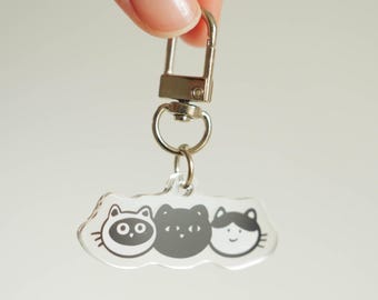 May include: A clear acrylic keychain featuring three cartoon cat faces in black and white. The keychain has a silver-colored clasp and ring for attaching to keys or bags. The cat faces have different expressions.
