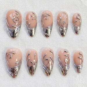 May include: Set of ten almond-shaped press-on nails. Each nail features a nude base with a silver metallic tip and a swirling, ornate design. The nails are arranged on a white, textured surface.