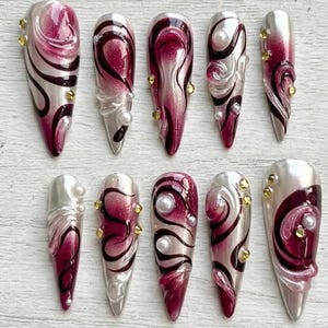 May include: A collection of ten long, stiletto-shaped false nails. Each nail is painted with a silver base, burgundy swirls, and black accents. Some nails feature pearls and gold-coloured rhinestones for added detail.