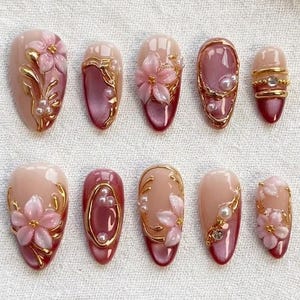 Op de afbeelding: Een set van tien kunstnagels met een bloemenontwerp. De nagels hebben een verloop van lichtroze naar dieprood, versierd met gouden accenten, parels en roze bloemversieringen. De nagels zijn ovaal van vorm.