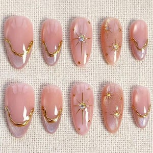 May include: Set of ten artificial nails in various sizes. The nails are oval-shaped with a light pink base and gold accents. Some nails feature a gold starburst design with a central gem, while others have a gold border.