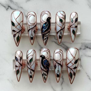 Puede incluir: Juego de diez uñas postizas largas en forma de almendra. Cada uña presenta una base nacarada con contornos color cobre. Los diseños incluyen lunas crecientes, pájaros negros, hojas y pequeñas botellas, con acentos verdes y azules.