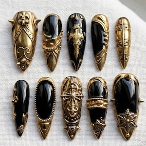 May include: An assortment of ten ornate, gold-colored nail art designs. Each nail features a black, glossy base with intricate gold embellishments, including spikes, crosses, and floral patterns. The nails are arranged on a white surface.