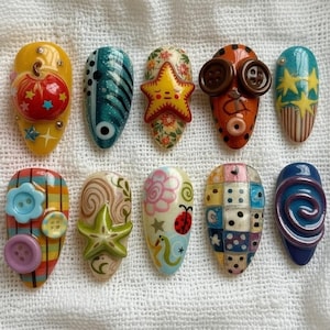 May include: A collection of ten uniquely designed artificial nails. Each nail features a different design, including a pumpkin, a starfish, buttons, and geometric patterns. The nails are painted in various colors, including yellow, blue, orange, and green.