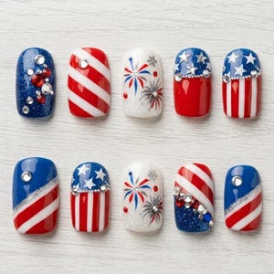Könnte beinhalten: Ein Set aus zehn künstlichen Nägeln mit patriotischen Designs. Die Nägel zeigen rote, weiße und blaue Farben mit Streifen, Sternen und Feuerwerksmustern. Einige Nägel sind mit Strasssteinen verziert.