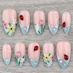 May include: A collection of ten false nails, each with a light pink base and a light blue tip. The nails are decorated with floral designs, ladybugs, and water droplet details. The oval-shaped nails are displayed on a wooden surface.