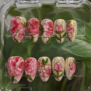 May include: A set of ten artificial nails with a floral design. Each nail features a pink rose design with gold outlines and green leaves, set against a pearlescent background. The nails are displayed in a clear plastic case.