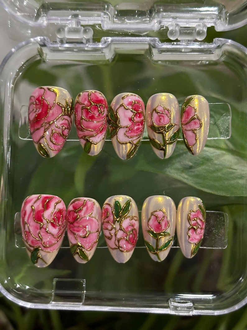 May include: A set of ten artificial nails with a floral design. Each nail features a pink rose design with gold outlines and green leaves, set against a pearlescent background. The nails are displayed in a clear plastic case.