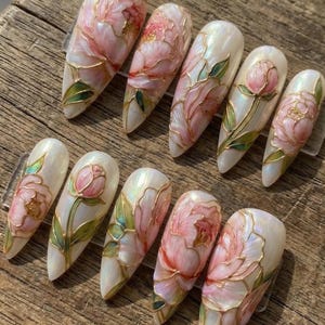 May include: A set of ten artificial nails with a pearlescent base, each adorned with a detailed floral design. The flowers are primarily pink with gold outlines and green leaves, creating an elegant and artistic look.