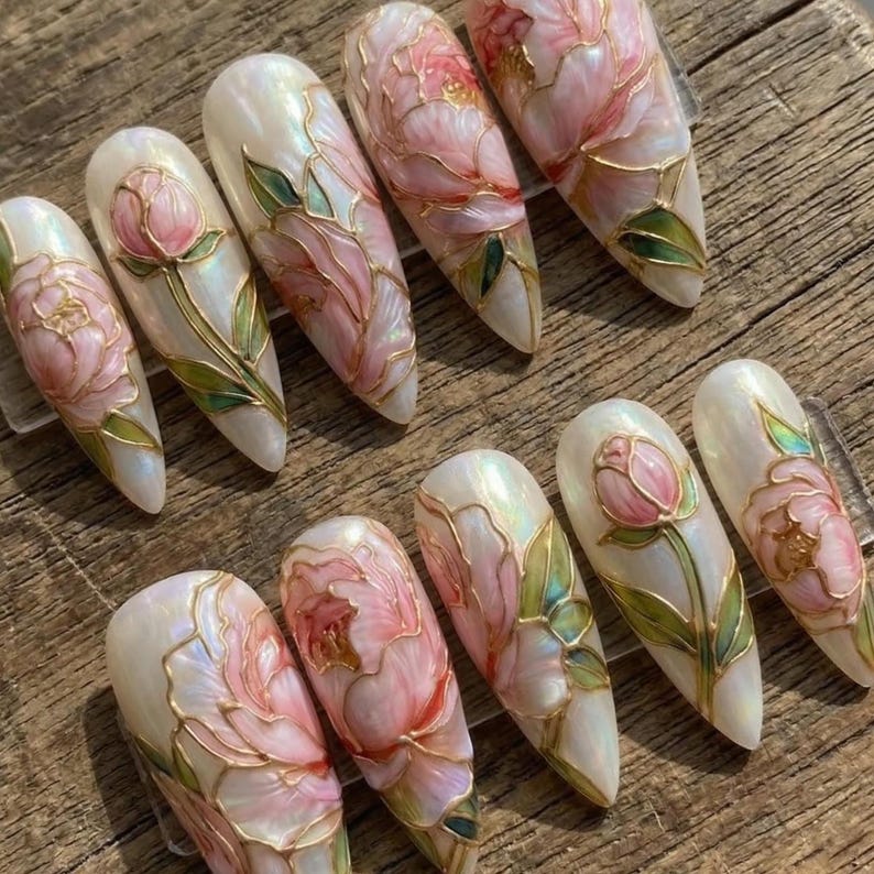 May include: A set of ten almond-shaped artificial nails. Each nail features a pearlescent base with a hand-painted floral design in pink, green, and gold. The flowers are detailed with gold outlines and green leaves.