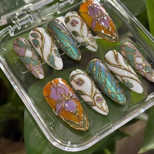 May include: A set of ten artificial nails in a clear plastic case. The nails feature various designs, including a purple flower on an orange background, iridescent green, white with gold filigree, and a pink and green diamond pattern. The nails are oval-shaped.