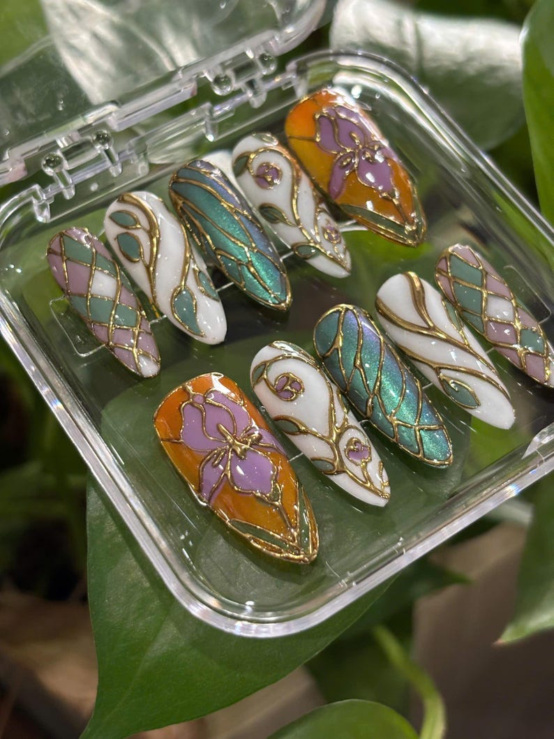 May include: A set of ten artificial nails in a clear plastic case. The nails feature various designs, including a purple flower on an orange background, iridescent green, white with gold filigree, and a pink and green diamond pattern. The nails are oval-shaped.