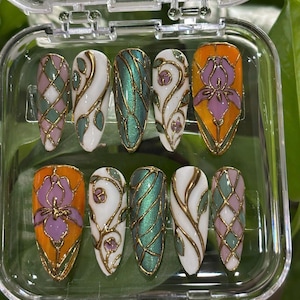 May include: A set of ten artificial nails with a variety of designs. The nails feature a mix of colors, including white, orange, green, and purple, with gold accents. Designs include floral patterns, geometric shapes, and abstract swirls, all encased in a clear plastic case.