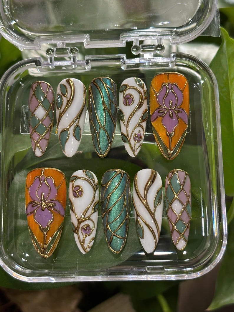 May include: A set of ten artificial nails with a variety of designs. The nails feature a mix of colors, including white, orange, green, and purple, with gold accents. Designs include floral patterns, geometric shapes, and abstract swirls, all encased in a clear plastic case.