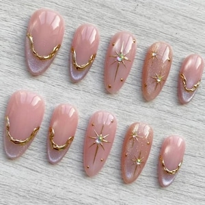 May include: A set of ten artificial nails in various sizes. The nails are primarily pink with gold accents. Some nails feature gold starburst designs with rhinestones, while others have a gold trim along the base. The nails have a glossy finish.