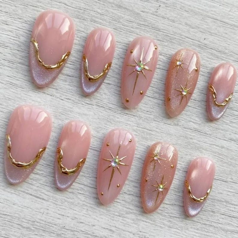 May include: A set of ten artificial nails in various sizes. The nails are primarily pink with gold accents. Some nails feature gold starburst designs with rhinestones, while others have a gold trim along the base. The nails have a glossy finish.