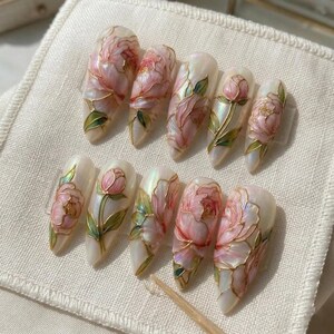 May include: A set of ten artificial nails with a floral design. Each nail features a pearlescent base with pink peony flowers, green leaves, and gold outlines. The nails are arranged on a white fabric surface.