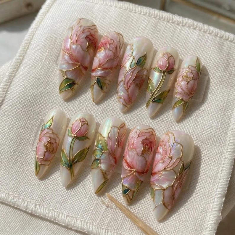 May include: A set of ten artificial nails with a floral design. Each nail features a pearlescent base with pink peony flowers, green leaves, and gold outlines. The nails are arranged on a white fabric surface.