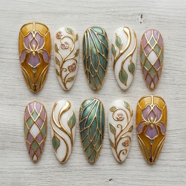 May include: A collection of ten almond-shaped nail art designs. Each nail features intricate gold outlines and details, with designs including floral patterns, geometric shapes, and iridescent green and purple accents. The nails are arranged on a light wood surface.