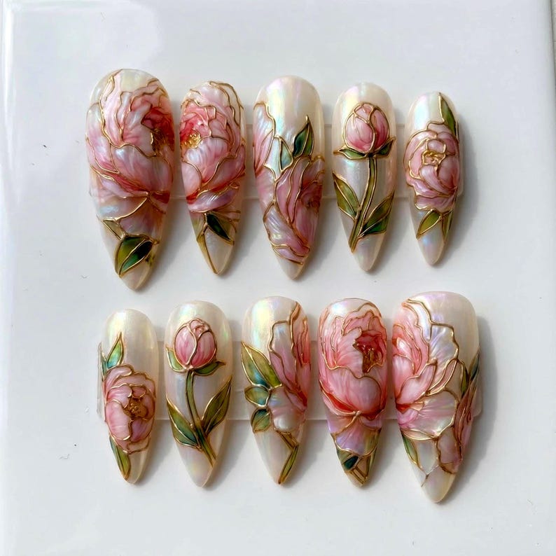May include: A set of ten almond-shaped artificial nails. Each nail features a pearlescent base with a hand-painted design of pink peonies with gold outlines and green leaves. The nails are arranged in two rows of five.