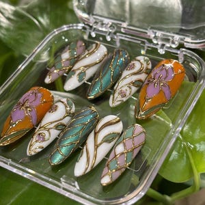 May include: A set of ten artificial nails in a clear plastic case. The nails feature various designs, including floral patterns, geometric shapes, and abstract swirls, with gold metallic accents. Colors include orange, white, green, and pink.