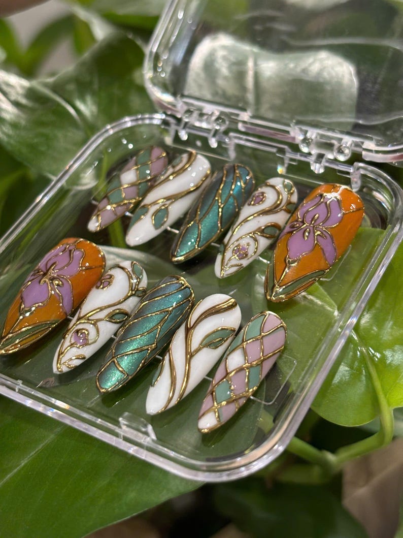 May include: A set of ten artificial nails in a clear plastic case. The nails feature various designs, including floral patterns, geometric shapes, and abstract swirls, with gold metallic accents. Colors include orange, white, green, and pink.